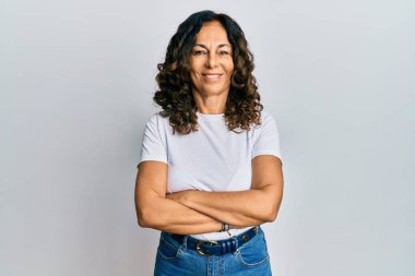 Orta yaşlı İspanyol kadın, günlük beyaz tişört giyip kollarını kavuşturup kameraya bakarken gülümsüyor. pozitif kişi. 