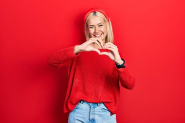 Güzel sarışın kadın, sıradan kırmızı bir süveter giyiyor. Sevgiyle gülümsüyor. Elleriyle kalp sembolü yapıyor. romantik kavram. 