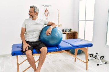 Orta yaşlı İspanyol bir adam ağrı kesici kliniğinde elinde pilates topu tutuyor. Yan tarafa bakıyor, profil pozu veriyor. Doğal bir yüzü ve kendine güvenen bir gülümsemesi var.. 