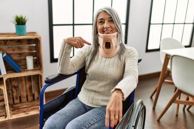 Orta yaşlı, gri saçlı, tekerlekli sandalyede oturan, tasmalı, mutlu ve gururlu birini işaret eden bir kadın. 