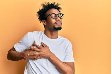 Sakallı, beyaz tişörtlü, gözlüklü, göğsünde elleri göğsünde, yüzünde minnet dolu bir jest ile gülümseyen Afrikalı genç bir Amerikalı. sağlık kavramı. 