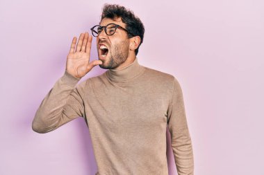 Balıkçı yaka kazak giyen, gözlüklü, sakallı, eli ağızda bağırıp çağıran yakışıklı bir adam. iletişim konsepti. 