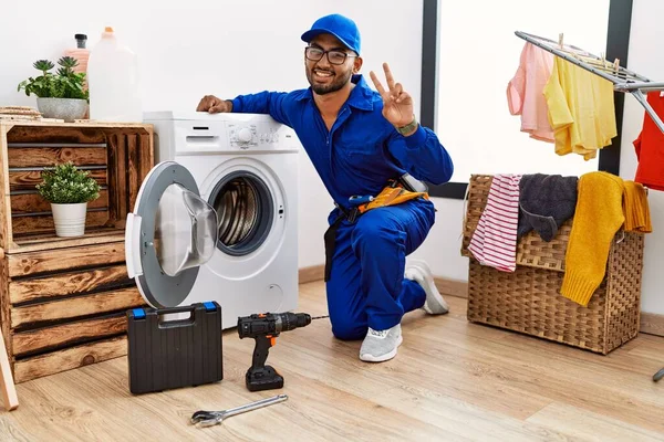 若いインドの技術者は 勝利のサインを行う指を示すカメラを見て笑みを浮かべて洗濯機に取り組んでいます — ストック写真