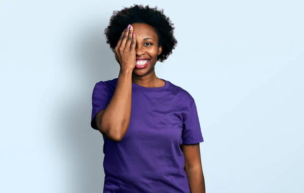 アフリカ系アメリカ人女性とアフロの髪を身に着けていますカジュアルパープルTシャツ手で一つの目をカバー 顔に自信の笑顔と驚きの感情 — ストック写真