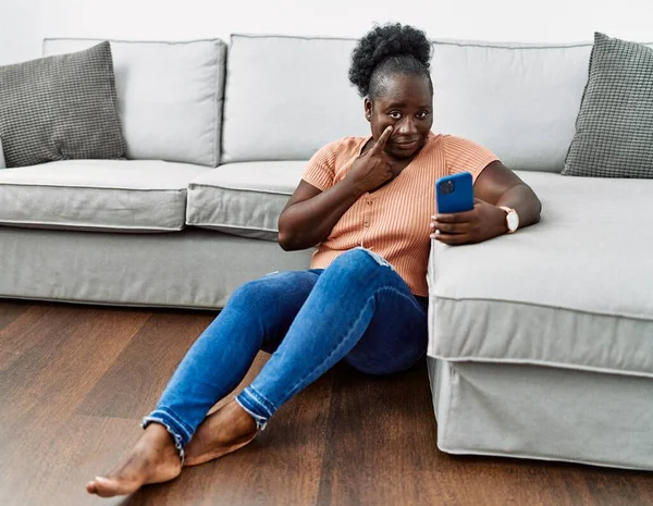 Молодая Африканская Женщина Используя Смартфон Сидя Дома Полу Указывая Глаз — стоковое фото