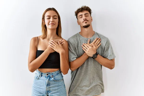 年轻漂亮的夫妇站在孤独的背景上 双手捂着胸膛 闭着眼睛 脸上挂着感激的手势 健康概念 — 图库照片