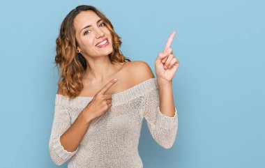 Güzel, beyaz bir kadın, günlük kıyafetler giymiş, gülümseyen ve kameraya bakıp iki elini ve parmağını yana doğru gösteren.. 