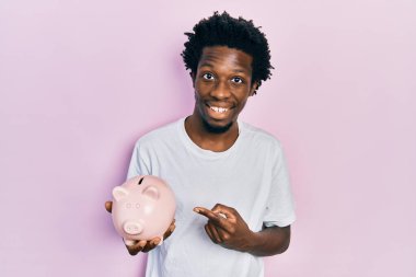 Genç Afrikalı Amerikalı domuz kumbarası tutan adam mutlu bir şekilde elini ve parmağını gösterdi. 