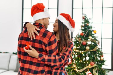 Mutlu gülümseyen ve ellerinde nişan yüzüğü tutan genç Latin çift Noel ağacının yanında duruyor..