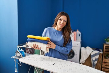 Ütü yaptıktan sonra katlanmış çamaşırları tutan genç esmer kadın pozitif ve mutlu görünüyor ve kendine güvenen bir gülümsemeyle dişlerini gösteriyor. 