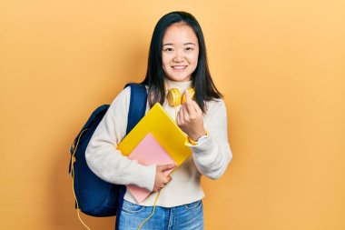 Öğrenci çantası ve kitapları tutan Çinli genç bir kız. Buraya el kol hareketi yaparak mutlu bir şekilde karşılamaya ve gülümsemeye davet ediyor. 