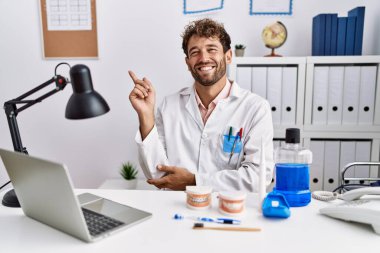 Tıbbi klinikte çalışan genç İspanyol dişçi yüzünde kocaman bir gülümsemeyle kameraya el ve parmak ile bakıyor.. 