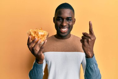 Genç Afrikalı Amerikalı adam elinde patates cipsiyle gülümsüyor bir fikir veya soru işareti ile mutlu bir yüzle, bir numara 