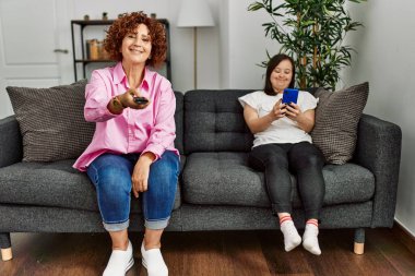 Olgun anne ve Down sendromlu kız evde televizyon izliyor ve akıllı telefon kullanıyor.