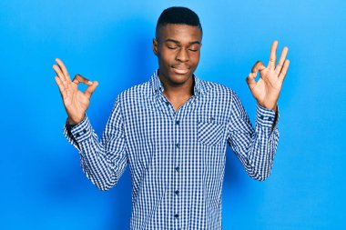 Genç Afro-Amerikan adam günlük kıyafetler giyip rahatlıyor ve gözleri kapalı gülümsüyor parmaklarıyla meditasyon hareketi yapıyor. yoga konsepti. 