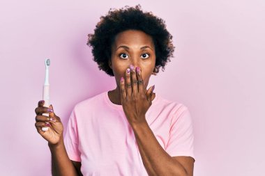 Genç Afro-Amerikalı kadın elinde elektrikli diş fırçası ile ağzını eliyle kapatıyor, şok olmuş ve hata yapmaktan korkuyor. şaşırmış ifade 