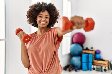 Genç Afrikalı Amerikalı kadın gülümsüyor spor merkezinde halterleri kullanarak kendine güven eğitimi alıyor