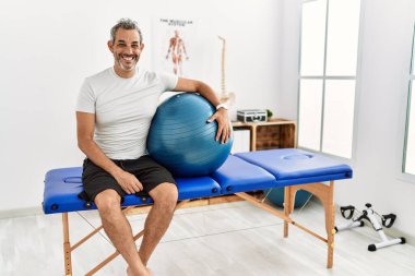 Orta yaşlı İspanyol bir adam ağrı kesici kliniğinde elinde mutlu ve havalı bir gülümsemeyle pilates topu tutuyor. şanslı kişi. 
