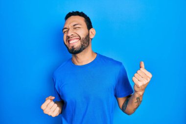 Sakallı, sıradan mavi tişörtlü İspanyol bir adam mutlu ve heyecanlı bir şekilde kolları havada, gülümseyen ve başarı için bağıran bir jest yapıyor. kutlama konsepti. 