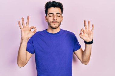 Günlük tişört giyen genç İspanyol adam rahatlamış ve gözleri kapalı gülümsüyor parmaklarıyla meditasyon hareketi yapıyor. yoga konsepti. 