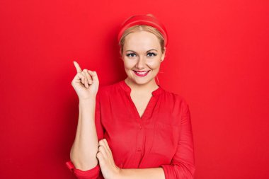 Sıradan kırmızı tişört giyen genç sarışın kadın gülümsüyor mutlu bir şekilde elini ve parmağını yana doğru gösteriyor. 