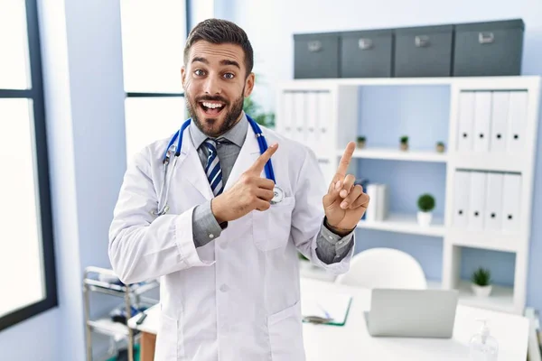 一个身穿医生制服 头戴听诊器 面带微笑的英俊男子站在诊所里 一边看着摄像机 一边用双手和手指指向旁边 — 图库照片