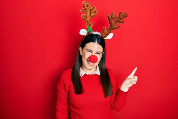 顔に大きな笑顔で鹿のクリスマスの帽子と赤い鼻を身に着けている若いヒスパニック系の女性 カメラを見て側に手指で指して — ストック写真