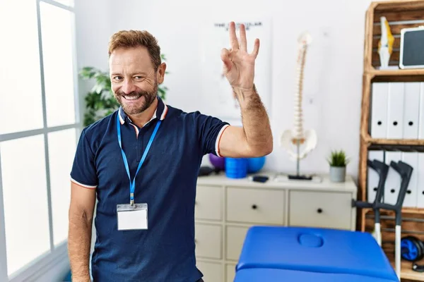 中年の理学療法士は 手や指でOkサインをして笑顔で痛み回復クリニックで働いています 成功した表現 — ストック写真