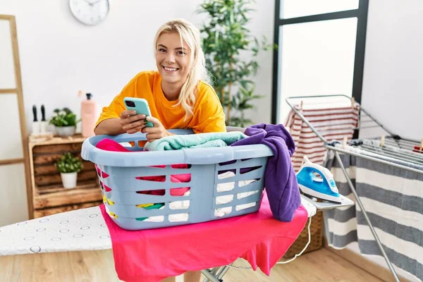 年轻的金发姑娘用智能手机靠在篮子上 家里有干净的衣服 — 图库照片