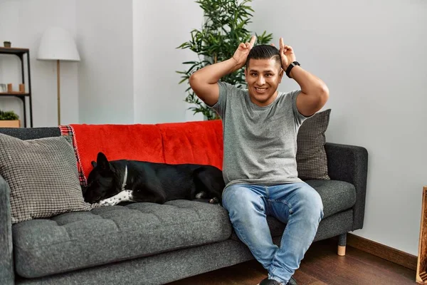 年轻的拉丁男人和狗坐在家里的沙发上 摆出一副滑稽而疯狂的样子 头上拿着手指 就像兔子的耳朵一样 开心地微笑着 — 图库照片