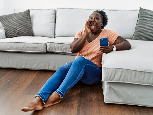 年轻的非洲女人用智能手机坐在家里的地板上大声喊着 手拉手地大声尖叫着 传播概念 — 图库照片