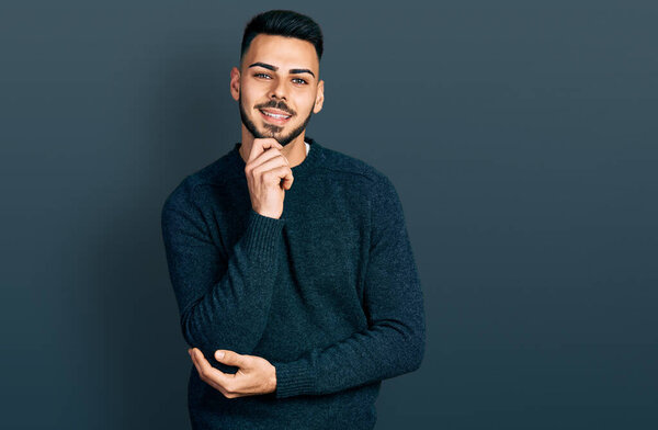 Young hispanic man with beard wearing casual winter sweater smiling looking confident at the camera with crossed arms and hand on chin. thinking positive. 