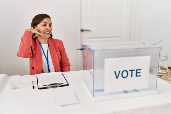 在政治选举中 美丽的中年惊慌失措的女人坐在投票站边投票边微笑着 手指手画脚地做着电话手势 就像在讲电话一样 交流概念 — 图库照片