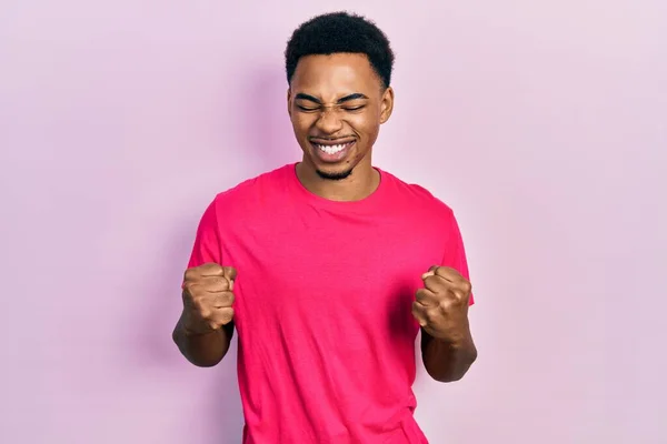 若いアフリカ系アメリカ人の男性が腕を上げて成功に興奮し 目を閉じて勝利笑顔を祝うカウアキャストシャツを着て 勝者のコンセプト — ストック写真