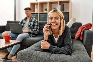Genç beyaz çift dizüstü bilgisayar kullanarak mutlu bir şekilde gülümsüyor ve evdeki akıllı telefondan konuşuyorlar..