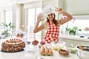 Güzel, genç, esmer pasta şefi kadın mutfakta hamur işleri pişiriyor. Sekiz numaralı parmaklarıyla işaret ediyor ve gülümsüyor. Kendinden emin ve mutlu.. 