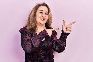 Orta yaşlı beyaz kadın günlük kıyafetler giyiyor gülümsüyor ve kameraya bakıyor iki eliyle ve parmaklarıyla kenarı işaret ediyor. 