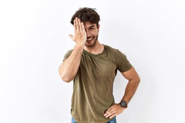 Hispanik adam, izole edilmiş beyaz arka planda duruyor. Bir gözünü eliyle kapatıyor. Yüzünde kendine güvenen bir gülümseme ve sürpriz bir duygu var.. 
