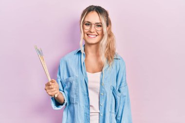 Güzel sarışın kadın elinde boya fırçaları tutarken pozitif ve mutlu görünüyor ve gülümsüyor. Kendine güvenen bir gülümsemeyle dişlerini gösteriyor. 
