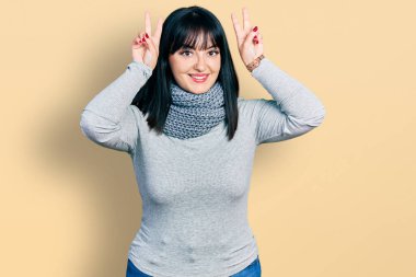 Genç Hispanik artı cüsseli bir kadın komik poz veriyor ve deli parmakları tavşan kulağı gibi, neşeli gülümsüyor. 