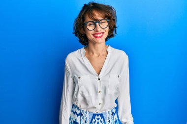 Genç esmer kadın günlük kıyafetler ve gözlük takıyor ve yüzünde mutlu ve havalı bir gülümseme var. şanslı kişi. 