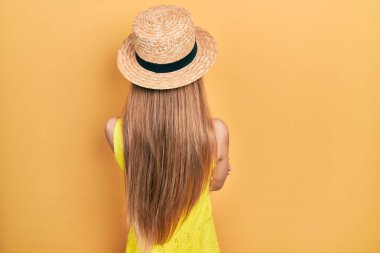 Sarışın, yaz şapkası takan genç kız kollarını kavuşturup arkasına bakıyor. 