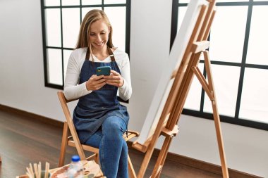 Beyaz kadın sanat stüdyosunda akıllı telefon kullanarak gülümsüyor.