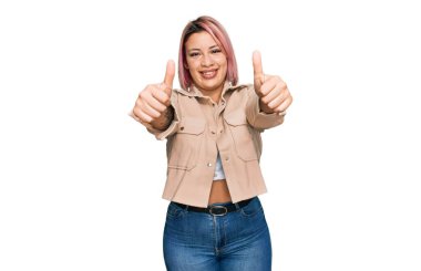 Pembe saçlı İspanyol bir kadın, günlük kıyafetlerini giyiyor. Elleriyle olumlu bir jest yapıyor. Gülümsüyor ve başarı için seviniyor. kazanan jest. 