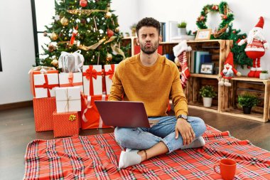 Arap genç adam dizüstü bilgisayar kullanıyor Noel ağacının yanında oturuyor dudaklarıyla balık suratı yapıyor, çılgınca ve komik hareketler yapıyor. Komik ifade. 