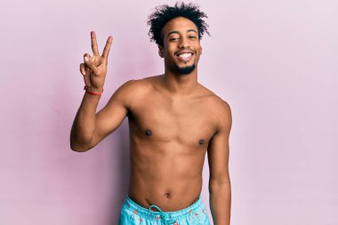 Sakallı, mayo giymiş, iki numaralı parmaklarıyla gösteriş yapan, kendinden emin ve mutlu genç Afrikalı Amerikalı bir adam.. 
