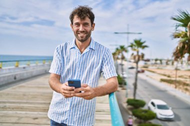 Genç İspanyol adam güvertede akıllı telefon kullanarak mutlu bir şekilde gülümsüyor..