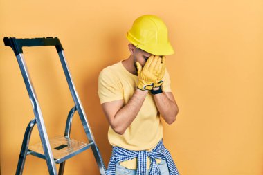 İnşaat merdivenlerinde sakallı yakışıklı bir adam. Ağlarken yüzünü ellerle kapatan askılı bir şapka takıyor. depresyon kavramı. 