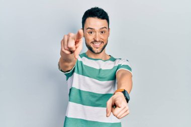 Gündelik kıyafetler giyen genç İspanyol adam seni ve kamerayı parmaklarıyla göstererek mutlu ve mutlu gülümsüyor. 