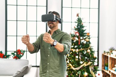 VR gözlük takan genç İspanyol adam Noel ağacının yanında duruyor.